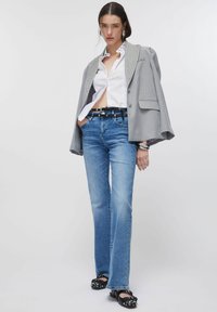 Blazer à carreaux gris avec accents cloutés, chemise blanche, jean évasé bleu clair, et chaussures noires ornées. Textures lisses partout.