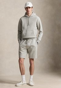 Homme portant un sweat à capuche gris clair, un short assorti, une casquette blanche, des chaussettes blanches et des baskets blanches, debout les mains dans les poches.