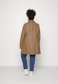 Brown coat with a tailored design, long sleeves, and a notched collar, worn over blue pants and white sneakers. Smooth texture.