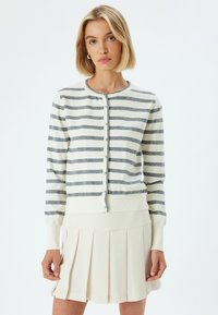 Biała kardiganka z szarymi poziomymi paskami, z okrągłym dekoltem, zapięciem na guziki i prążkowanymi mankietami. Nosi ją na plisowanej beżowej spódnicy.