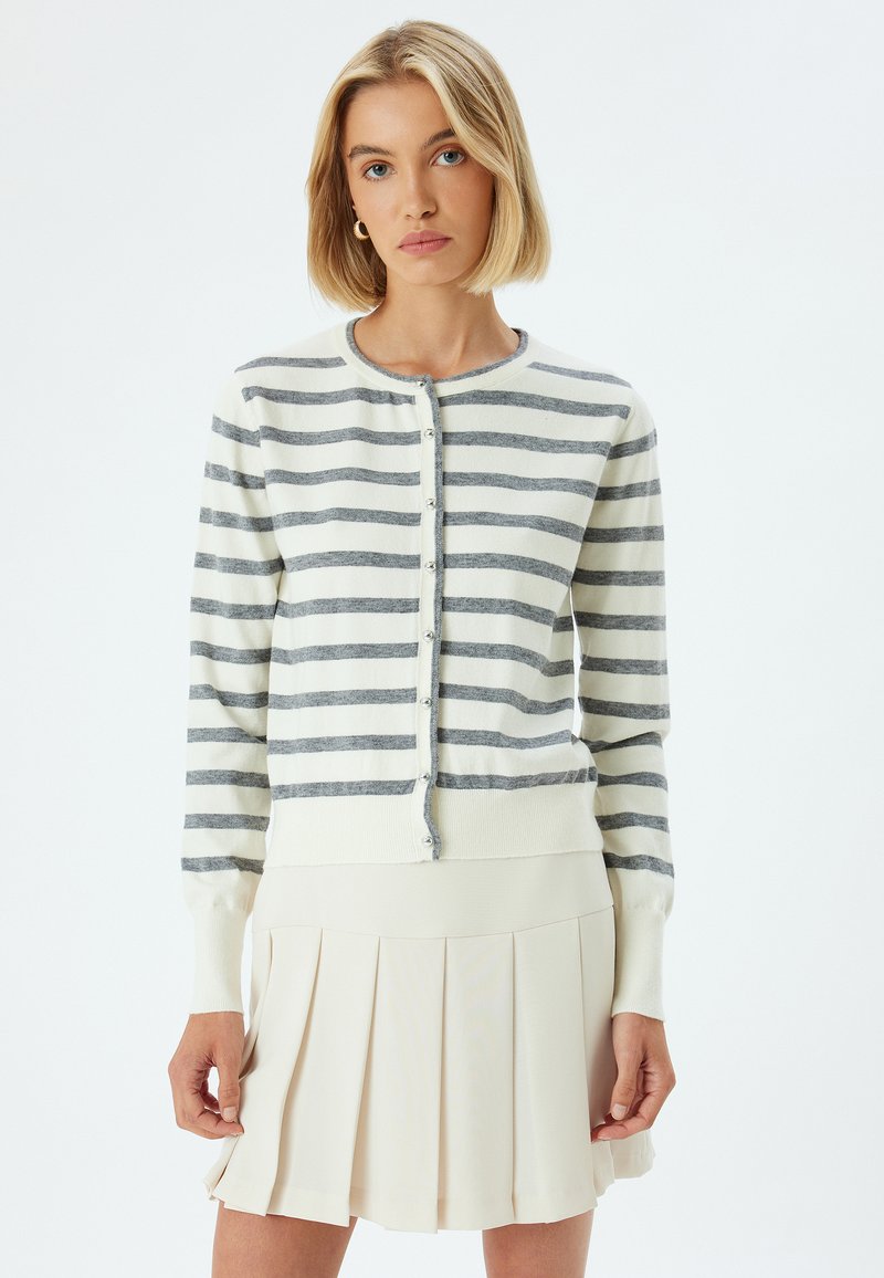 Biała kardiganka z szarymi poziomymi paskami, z okrągłym dekoltem, zapięciem na guziki i prążkowanymi mankietami. Nosi ją na plisowanej beżowej spódnicy.