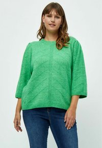 Grüner Strickpullover mit strukturiertem Zickzackmuster, weiten Ärmeln und rundem Halsausschnitt, kombiniert mit blauen Jeans.