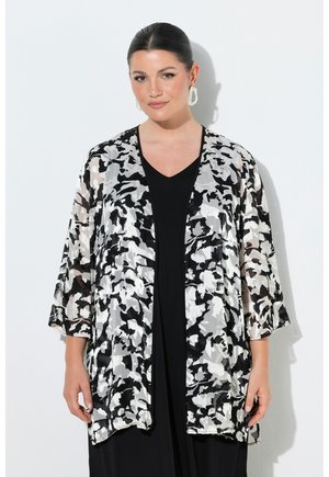 Frau mit dunklem Haar in einem niedrigen Dutt, die große weiße Ohrringe, ein schwarzes Kleid und eine lockere, schwarz-weiß gemusterte, offene Strickjacke trägt.