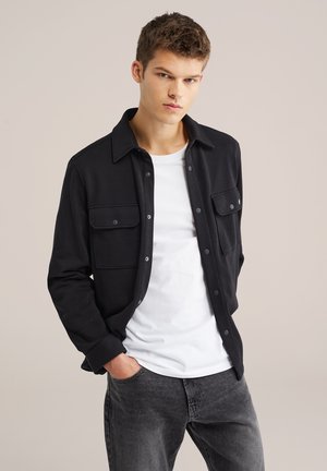 Jonge man die een zwart overhemd draagt over een wit T-shirt en grijze jeans, staand met één hand in zijn zak tegen een effen achtergrond.