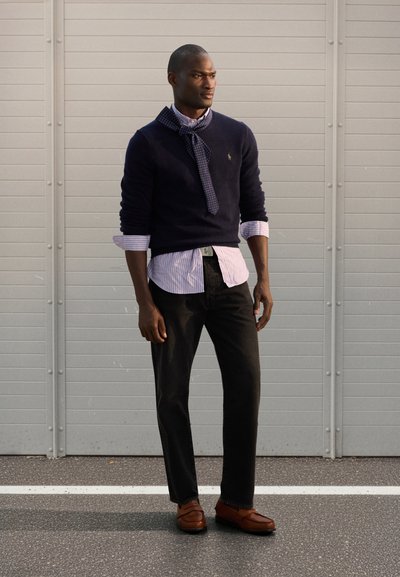 Ναυτικό πουλόβερ πάνω από ένα γαλάζιο ριγωτό πουκάμισο, σε συνδυασμό με σκούρο τζιν και καφέ μοκασίνια. Το κασκόλ έχει ναυτικά πουά. Ουδέτερο φόντο.