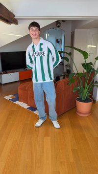 Zöld-fehér csíkos mez „F.C. HOUSE” felirattal, világoskék farmernadrág, fehér tornacipő, egy világos szobában áll, növényekkel és bútorokkal.