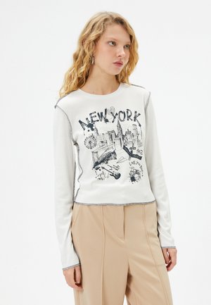 T-shirt bianco a maniche lunghe con grafica "NEW YORK", dettagli neri e texture a coste. Abbinato a pantaloni beige, con vestibilità dritta.
