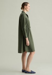 Grünes Hemdkleid mit Kragen, langen Ärmeln und gerafftem Taillenbereich. Der Stoff hat ein feines Muster. Kombiniert mit blauen, gemusterten Ballerinas.