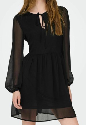 Woman wearing a black sheer long-sleeve dress with a tie at the neckline and gathered waist, standing against a white background.