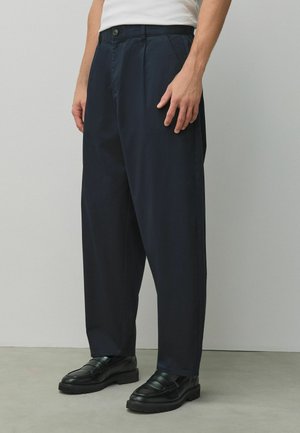 Man wearing white shirt, navy loose-fit trousers, and black leather loafers, standing against a plain gray wall.