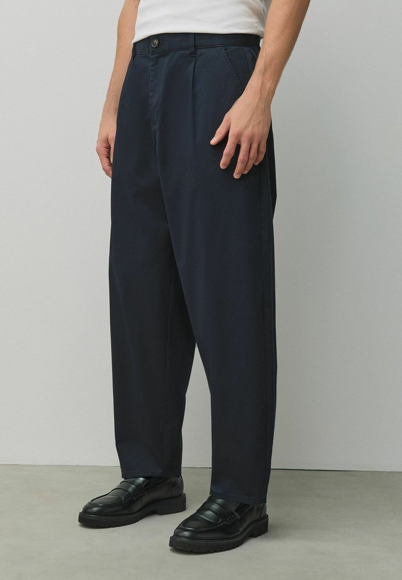 Mann trägt weißes Hemd, marineblaue locker sitzende Hose und schwarze Ledermokassins, steht vor einer schlichten grauen Wand.