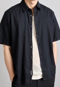 Zwarte shirt met korte mouwen, een kraag, knopen aan de voorkant en een losse pasvorm, gelaagd over een licht mesh top.