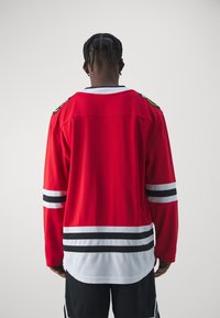 Maglia da hockey rossa con maniche lunghe, strisce bianche e nere sulle maniche e sull'orlo, con toppe con logo sulle spalle, realizzata in tessuto a rete.