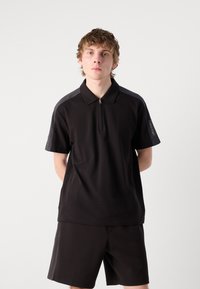 Čierne polo tričko na zips s krátkymi rukávmi, ktoré má tmavšie ramenné akcenty a jemný detail loga na rukáve, v kombinácii s čiernymi šortkami.