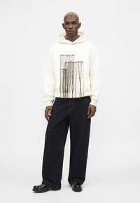 Αρσενικό μοντέλο με κοντά ξανθά μαλλιά που φοράει μια κρεμ φούτερ με σχέδιο μαύρου κορδονιού και κείμενο, φαρδιά μαύρα παντελόνια και μαύρα παπούτσια, όρθιος.