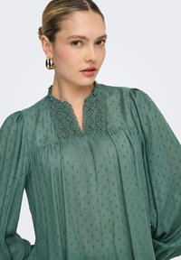 Γυναίκα με ξανθά μαλλιά σε χαμηλό κότσο, φορώντας πράσινη υφασμάτινη μπλούζα με δαντελένιες λεπτομέρειες και χρυσούς κρίκους σκουλαρίκια, σε ανοιχτό φόντο.
