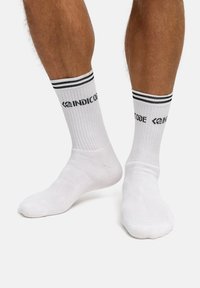 Białe bawełniane skarpetki o prążkowanej fakturze, z czarnymi paskami na górze i czarnym logo "INDICODE" blisko mankietu.