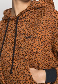 Felpa arancione con stampa leopardata nera. Caratterizzata da un cappuccio con coulisse, tasca frontale e polsini a coste. Il materiale è morbido e testurizzato.