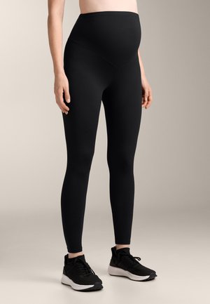 Schwarze Schwangerschaftsleggings mit hoher Taille und schmalem Schnitt, aus dehnbarem Stoff, kombiniert mit schwarzen Sportschuhen.