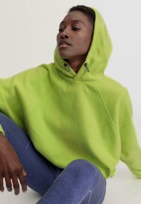Limettengrüner Fleece-Hoodie mit Kordelzugkapuze, Raglanärmeln und weicher Textur. Kombiniert mit dunkelblauen, strukturierten Leggings.