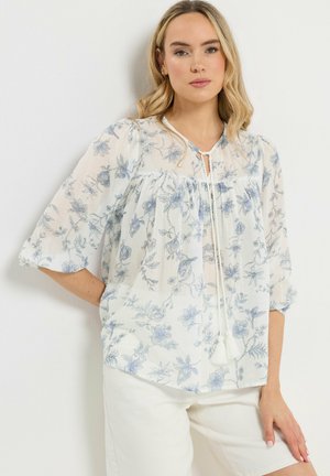 Kvinde iført en hvid gennemsigtig bluse med blåt blomsterprint og kvaster, kombineret med hvide bukser, stående op ad en ensfarvet hvid væg.