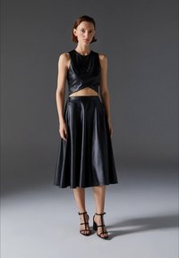 Top corto in pelle nera con un design attorcigliato e una gonna coordinata fluida, lunghezza ginocchio. L'outfit presenta linee eleganti e sandali neri minimalisti.