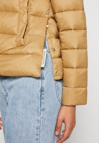 Veste matelassée beige avec boutons-pression portée sur un jean en denim bleu clair, montrant une étiquette en tissu blanche et la main droite d'une personne détendue sur le côté.