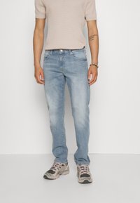 Vaqueros de mezclilla azul claro con corte recto, lavado desvanecido y diseño de cinco bolsillos. Combinados con una camiseta de punto acanalado beige y zapatillas deportivas.