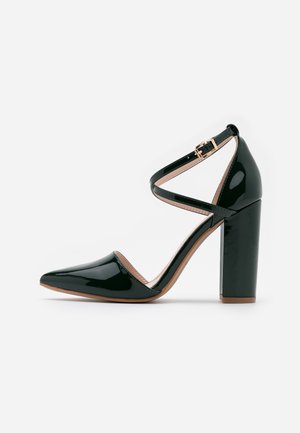 Zapato de tacón alto de charol negro con punta puntiaguda, correa en el tobillo y tacón ancho grueso. Presenta un diseño de tiras cruzadas y un interior en tono nude.