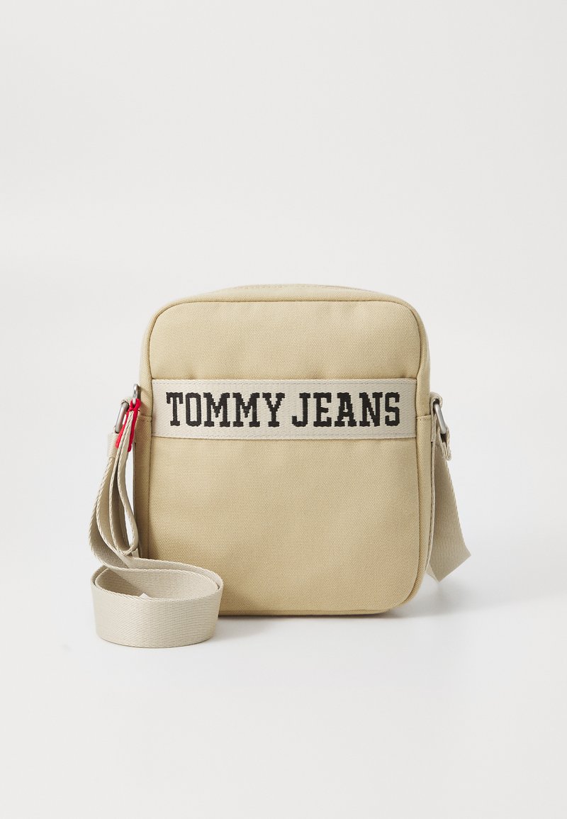 Beige lerretscrossbody veske med svart "TOMMY JEANS" tekst, justerbar stropp og glidelås. Har et enkelt rektangulært design.