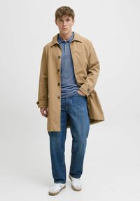 Beige Trenchcoat mit Kragen und Knöpfen, getragen über einem blauen Poloshirt. Kombiniert mit blauen Jeans und weißen Sneakers mit beigen Akzenten.