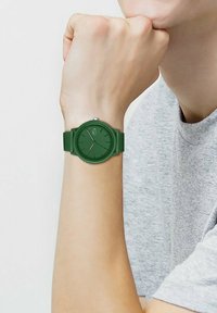Montre en silicone verte avec un cadran rond et un fond texturé. Présente des marqueurs d'heure minimalistes et un petit logo au centre.