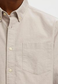 Φωτεινό μπεζ πουκάμισο με κουμπιά, γιακά και μία τσέπη στο στήθος, με περλέ κουμπιά. Το ύφασμα φαίνεται λείο με διακριτική υφή.