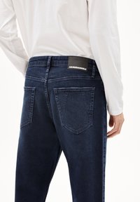 Donkerblauwe jeans met een strak ontwerp, met een rechthoekig zwart label met witte tekst op de achterkant van de taille en twee achterzakken.