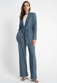 Jasnoniebieska dopasowana marynarka z wiązaniem w talii, w komplecie ze szerokimi spodniami. Gładka tekstura tkaniny; posiada dekoracyjny broszkę.