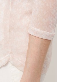 Camicia leggera rosa chiaro, trasparente, con un delicato motivo floreale, dotata di maniche a tre quarti e un orlo semplice. Tessuto morbido.