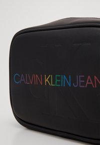 Černá obdéln�íková kosmetická taštička s reliéfním nápisem "CALVIN KLEIN JEANS" v gradientním duhovém barevném schématu na jedné straně.