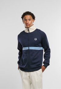 Mornarska zip-jakna s visokim kremastim ovratnikom, svetlo modrimi vodoravnimi črtami in stranskimi žepi; ima logotip na levi prsi.