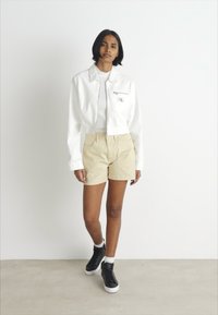 Giacca bianca corta con tasca con zip, abbinata a pantaloni corti beige. Sneakers nere con suola bianca completano l'outfit. Sfondo neutro.