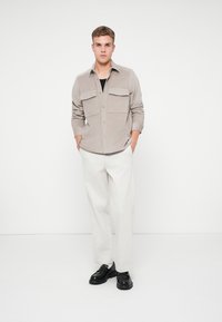 Camisa de botón en gris claro con dos bolsillos en el pecho, combinada con pantalones anchos blancos y zapatos negros, posicionada contra un fondo sencillo.