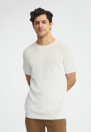 Junger Mann mit dunklem lockigem Haar, der einen weißen kurzärmligen Strickpullover und braune Hose trägt, mit hinter dem Rücken verschränkten Händen stehend.