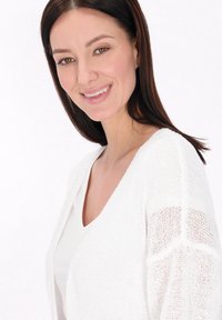Biely pletený kardigán s otvoreným predným dielom a textúrovaným vzorom, nosený cez biely top. Modelka má vlasy po plecia tmavej farby.