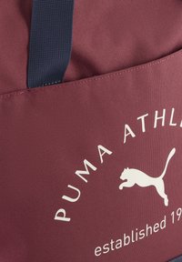 Burgundin väriä oleva kantokassi, jossa on teksturoitu kangas, siniset kahvat, valkoinen "PUMA ATHLETICS" -logo ja hyppivä puuma -grafiikka. Perustettu 1948 -yksityiskohta.