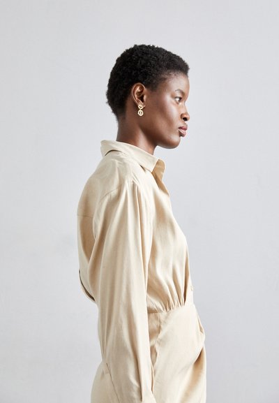 Camisa beige de botones con cuello estructurado y mangas abullonadas, que cuenta con una cinturilla elástica y está detallada con costuras mínimas.