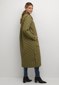 Olive zaļā quilted garā mētelis ar kapuci, kas aprīkots ar sānām kabatām un spiedpogu aizdari, kombinēts ar piegulošām zilām džinsām un melniem zamšāda zābakiem ar papēdi.