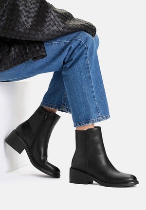 Osoba siedząca ze skrzyżowanymi nogami, nosząca czarne skórzane buty za kostkę, niebieskie dżinsy skrócone powyżej kostki oraz czarną plecioną kurtkę.