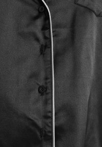 Schwarzes Hemd mit Knöpfen und glatter Textur, mit weißem Paspel entlang der Knopfleiste und einer einzelnen Brusttasche. Zwei sichtbare schwarze Knöpfe.