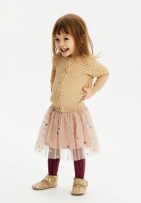 Beige kötött kardigán texturált részletekkel, rózsaszín tüll szoknya színes pöttyökkel, bordó harisnya és arany díszítésű cipők.