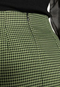 Gros plan sur un pantalon à motif pied-de-poule vert avec un haut noir, mettant en valeur la texture du tissu et un détail de fermeture éclair.