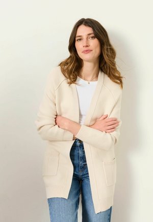 Woman with long wavy brown hair wearing a cream cardigan over a white shirt and blue jeans, standing with arms crossed against a plain background.